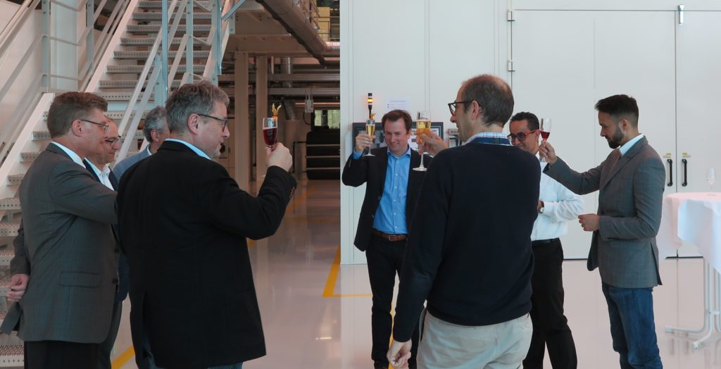 Group of men raising champagne glasses in toast while standing next to completed laser peening facility